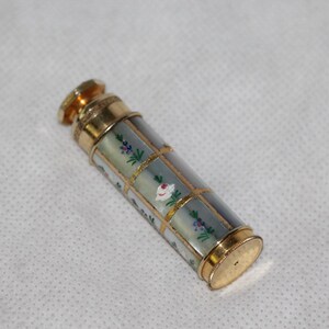 Pode incluir: Um frasco de perfume cilíndrico com uma estrutura de metal dourado em padrão de grade. O corpo consiste em painéis iridescentes decorados com pequenos motivos florais pintados à mão em rosa, verde e azul. Possui uma tampa e base douradas.