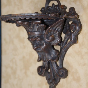May include: A dark brown, cast iron wall shelf with a decorative face and wing design. The shelf has a scalloped edge and is mounted on a wall. The shelf is approximately 6 inches wide.