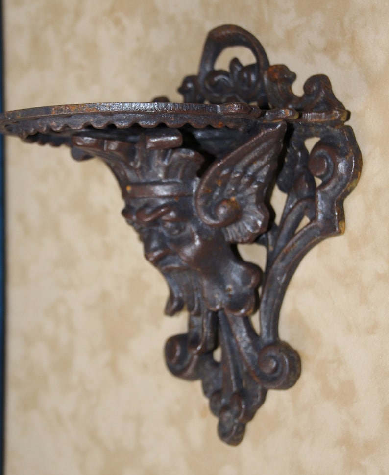 May include: A dark brown, cast iron wall shelf with a decorative face and wing design. The shelf has a scalloped edge and is mounted on a wall. The shelf is approximately 6 inches wide.