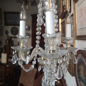 May include: A clear crystal candelabra with three candle holders. The candelabra features hanging crystal accents and a decorative base. The candle holders have white candle-shaped bulbs. The candelabra is on a brown surface.