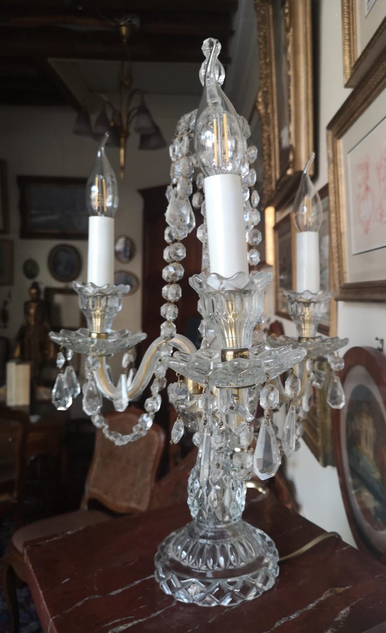 May include: A clear crystal candelabra with three candle holders. The candelabra features hanging crystal accents and a decorative base. The candle holders have white candle-shaped bulbs. The candelabra is on a brown surface.