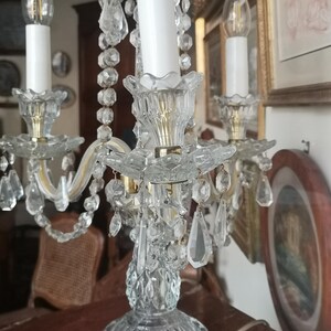 May include: A crystal candelabra with three candle holders. The candelabra has a clear, faceted base and is adorned with hanging crystal beads and teardrop-shaped crystals. The candle holders are gold-toned, and the candles are white.