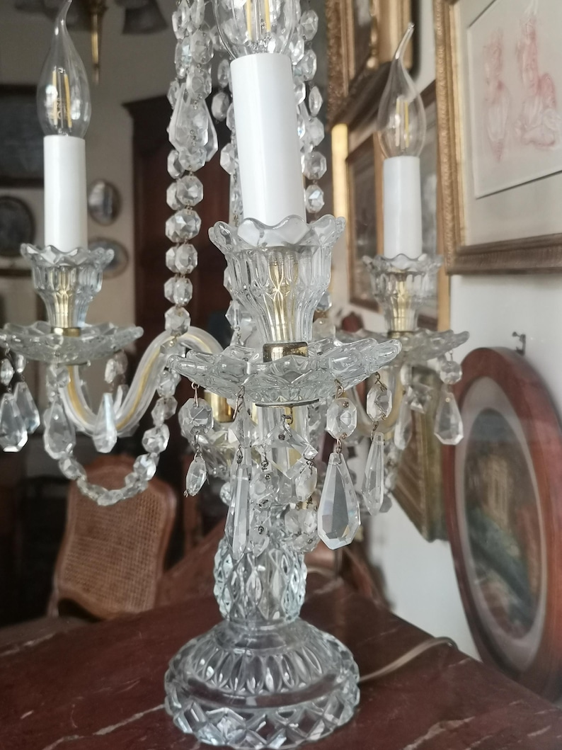May include: A crystal candelabra with three candle holders. The candelabra has a clear, faceted base and is adorned with hanging crystal beads and teardrop-shaped crystals. The candle holders are gold-toned, and the candles are white.