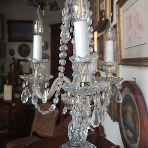 May include: A clear crystal chandelier-style lamp with three candle-shaped light bulbs. The lamp features ornate crystal accents, including hanging teardrop-shaped crystals. The base is also crystal, and the lamp sits on a dark red marble surface.