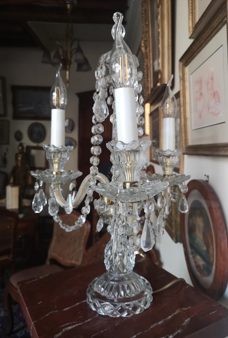 May include: A clear crystal chandelier-style lamp with three candle-shaped light bulbs. The lamp features ornate crystal accents, including hanging teardrop-shaped crystals. The base is also crystal, and the lamp sits on a dark red marble surface.