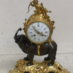 May include: An antique clock with a bronze elephant base and gold floral detailing. The clock face has black Roman numerals on a white background. A gold cherub with a bow and arrow is perched on top.
