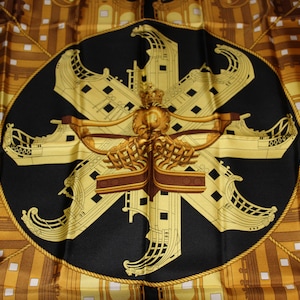 May include: A black silk scarf with a gold design of ships and a rope border. The ships are arranged in a starburst pattern.
