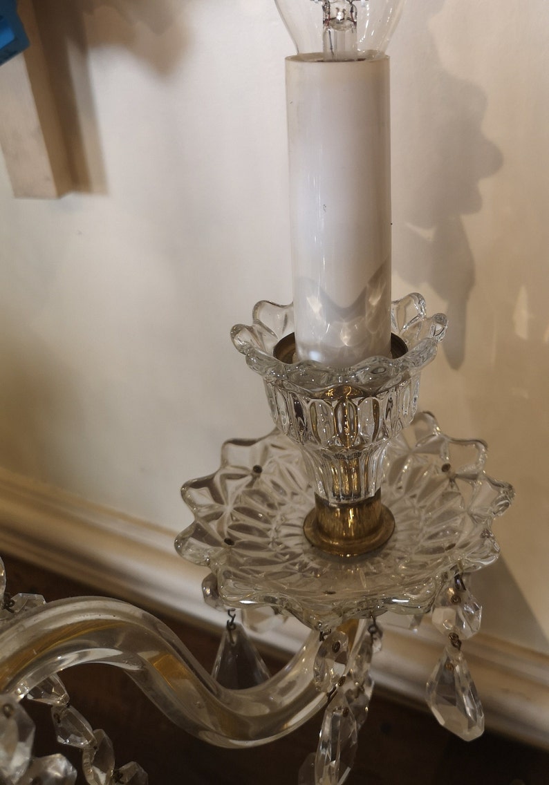 May include: A vintage crystal chandelier with a white candle-shaped light bulb. The chandelier has a clear, patterned glass base with a gold-coloured metal accent. Decorative crystal pendants hang from the base.