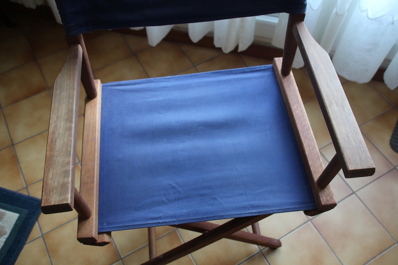 May include: A blue canvas director's chair with wooden arms and legs. The chair is folded and sitting on a tiled floor.