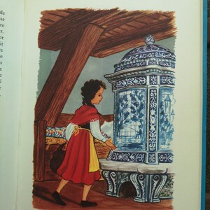 May include: A young girl in a red dress with a yellow sash stands in front of a blue and white tiled stove. The stove has intricate designs and a glass door. The girl is looking at the stove with a curious expression.