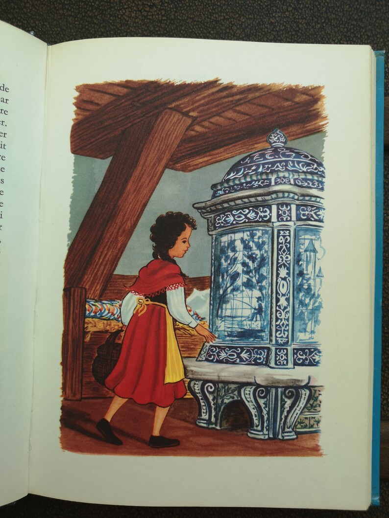 May include: A young girl in a red dress with a yellow sash stands in front of a blue and white tiled stove. The stove has intricate designs and a glass door. The girl is looking at the stove with a curious expression.