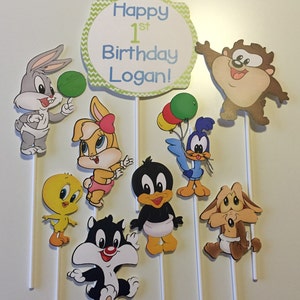 May include: A set of 9 baby Looney Tunes character cake toppers. The toppers feature Bugs Bunny, Tweety Bird, Sylvester, Daffy Duck, Lola Bunny, Tasmanian Devil, and Road Runner. The set also includes a sign that says "Happy 1st Birthday Logan!"