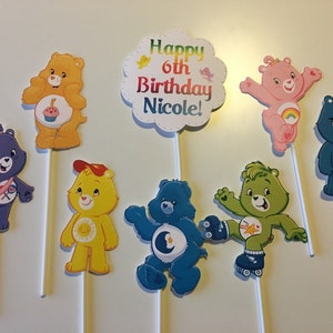 May include: Set of 7 Care Bear cake toppers. The toppers feature different Care Bears with their signature symbols. The topper with the cloud says "Happy 6th Birthday Nicole!"