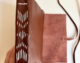 Könnte beinhalten: Ein braunes Lederjournal mit weißen Nähten entlang des Rückens. Das Journal ist geöffnet und zeigt ein leeres Innere und einen passenden braunen Lederriemen. Das Journal wird in einer Hand gehalten, um die Handwerkskunst und das Design zu präsentieren.