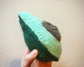 Könnte beinhalten: Ein gestricktes Avocado-Spielzeug mit einem grünen und braunen Farbschema. Die Avocado ist birnenförmig und hat einen Kern aus braunem Garn.