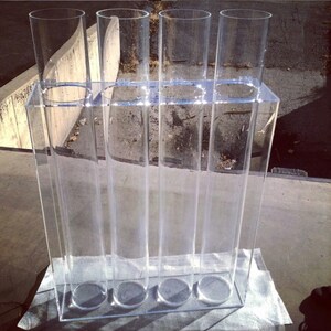 May include: A clear acrylic stand with five clear cylindrical vases. The stand has a rectangular base and is designed to hold the vases upright.