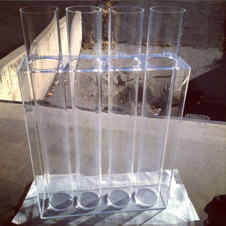 May include: A clear acrylic stand with five clear cylindrical vases. The stand has a rectangular base and is designed to hold the vases upright.