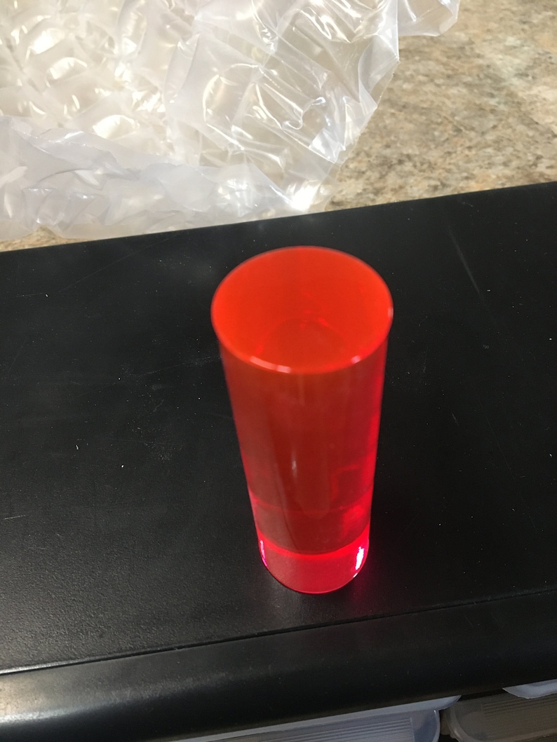 May include: A bright red shot glass made of clear plastic. The glass is empty and sitting on a black surface.