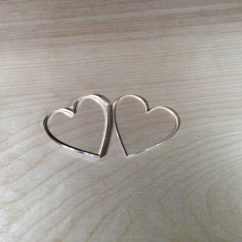 May include: Two clear acrylic heart shapes on a light brown wooden surface.