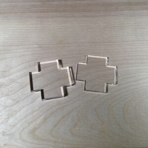 May include: Two clear plastic cross-shaped stencils on a light wood surface.