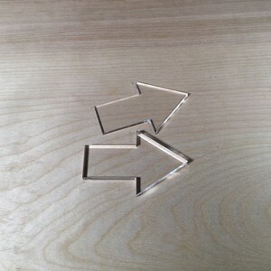 May include: Two clear plastic arrows pointing in opposite directions. The arrows are on a light brown wooden surface.