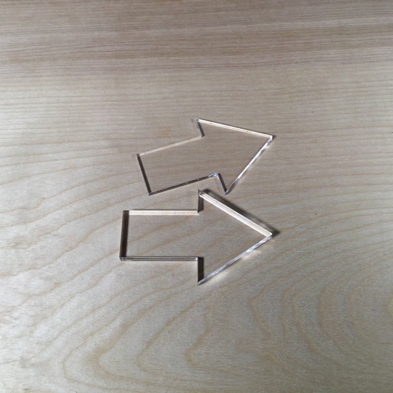 May include: Two clear plastic arrows pointing in opposite directions. The arrows are on a light brown wooden surface.