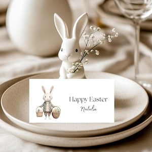 May include: A white ceramic bunny figurine holding a sprig of white flowers, placed on a layered plate setting. A card with the text "Happy Easter Natalie" is in front of the bunny. The setting is on a beige tablecloth.