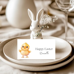 May include: Easter table setting featuring a white ceramic bunny holding flowers, a plate with a card that says "Happy Easter Danielle", and a watercolor chick illustration. The setting includes neutral-toned plates and glassware.