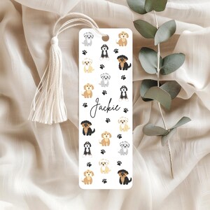May include: A white bookmark decorated with illustrations of different dog breeds and paw prints. The name "Jackie" is written in cursive. A cream tassel hangs from the top. A delightful accessory for book lovers.