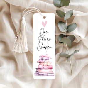 May include: A white bookmark with a tassel, featuring the text "One More Chapter" and a stack of watercolour books in shades of pink and purple. A small pink heart is above the text.