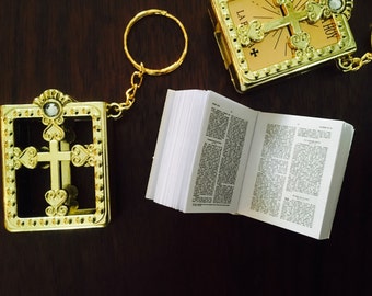 May include: A gold-colored, ornate, cross-shaped keychain with a small, clear crystal. The keychain is attached to a small, gold-colored, book-shaped charm. The book is open and has white pages with black text. The keychain is on a dark brown wooden surface.