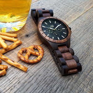 Op de afbeelding: Een donkerbruin houten polshorloge met een zwarte wijzerplaat en gouden accenten. Het horloge heeft een chronograaffunctie en het woord "Wild" staat op de wijzerplaat gedrukt.