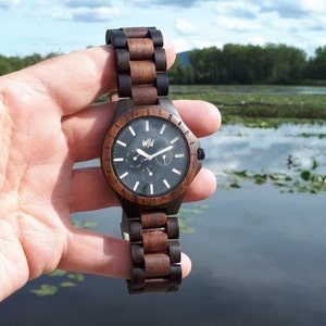 Op de afbeelding: Een bruin en zwart houten polshorloge met een zwarte wijzerplaat en witte wijzers. Het horloge heeft een datumvenster en een chronograaf. Het horloge wordt in een hand gehouden tegen een achtergrond van een meer en bomen.