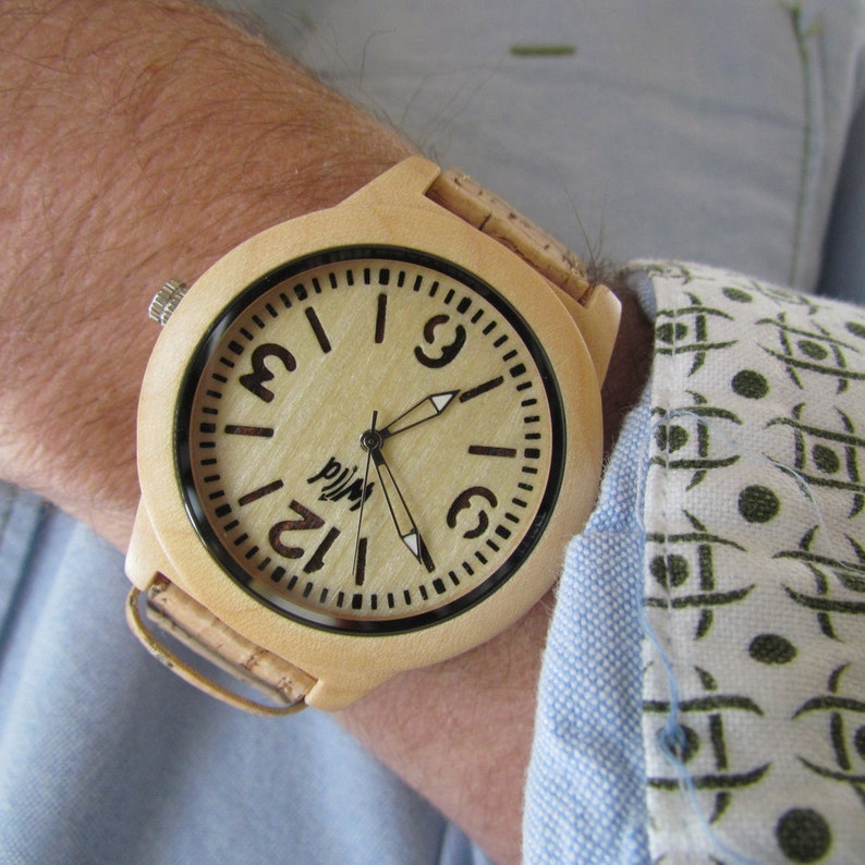 Puede incluir: Un reloj de pulsera de madera marr&oacute;n claro con una banda de corcho. La esfera del reloj es blanca con marcas negras y la palabra "Wild" impresa en ella.