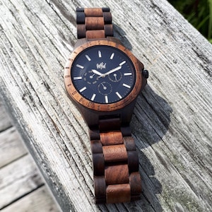 Op de afbeelding: Een bruin houten polshorloge met een zwarte wijzerplaat en witte wijzers. Het horloge heeft een chronograaffunctie en een datumscherm. Op de wijzerplaat van het horloge staat "Wild".