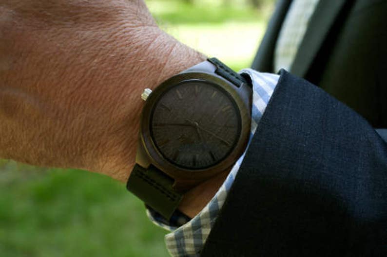 May include: A dark brown wooden wristwatch with a black leather band. The watch face has a dark wood grain pattern.