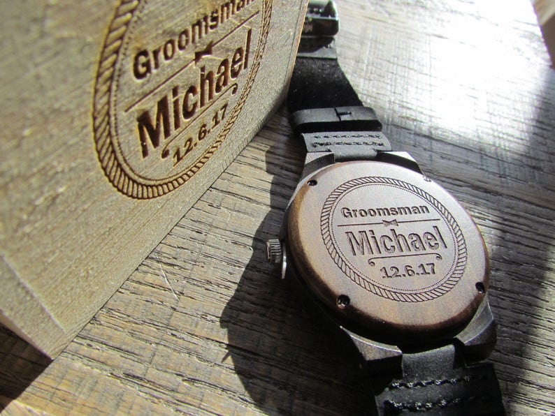May include: A wooden watch with a black leather band. The back of the watch is engraved with the text "Groomsman Michael 12.6.17".