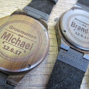 May include: Two wooden wristwatches with black leather bands. The watch faces are engraved with the words "Groomman" and the names "Michael" and "Brandon" along with the date "12.6.17".