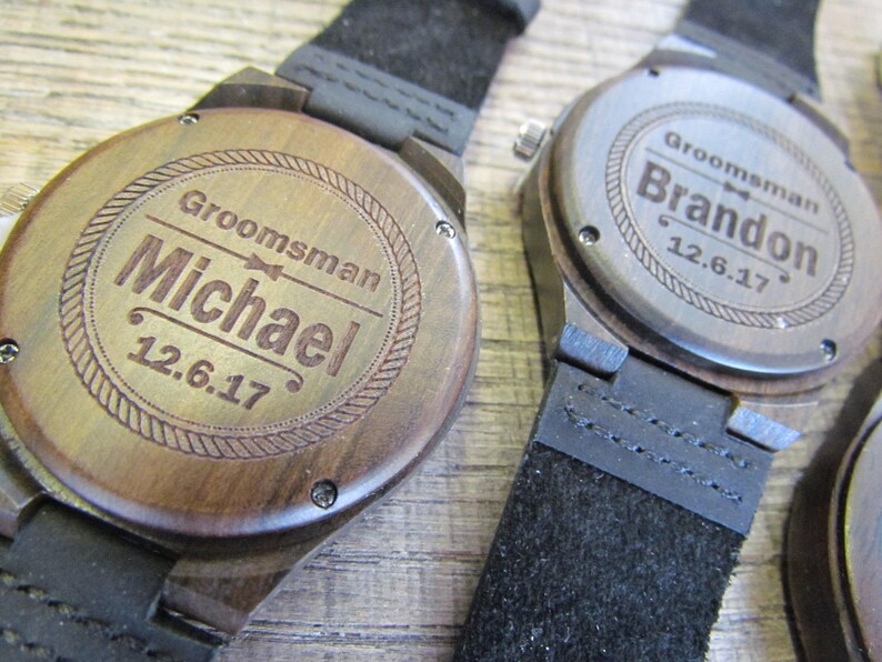 May include: Two wooden wristwatches with black leather bands. The watch faces are engraved with the words "Groomman" and the names "Michael" and "Brandon" along with the date "12.6.17".