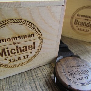 May include: Two wooden boxes with engraved text. The box on the left says "Groomsman Michael 12.6.17". The box on the right says "Groomsman Brandon 12.6.17". A wooden watch with a brown leather band is in the foreground. The watch face is engraved with "Groomsman Michael 12.6.17".