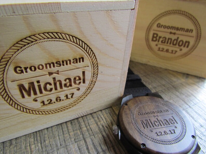 May include: Two wooden boxes with engraved text. The box on the left says "Groomsman Michael 12.6.17". The box on the right says "Groomsman Brandon 12.6.17". A wooden watch with a brown leather band is in the foreground. The watch face is engraved with "Groomsman Michael 12.6.17".