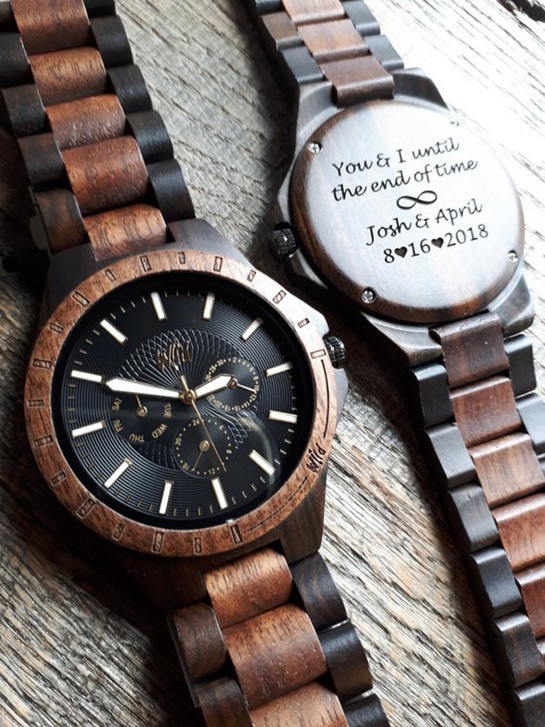 Op de afbeelding: Een houten polshorloge met een zwarte wijzerplaat en gouden accenten. Het horloge heeft een datumvenster, een dag van de week venster en een 24-uurs tijdvenster. De horlogeband is gemaakt van donker en licht hout. De achterkant van het horloge heeft een gepersonaliseerde inscriptie met de tekst "You & I until the end of time Josh & April 8 16 2018".