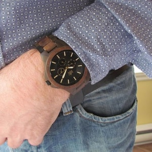 Op de afbeelding: Een bruin houten polshorloge met een zwarte wijzerplaat en zilveren wijzers. Het horloge heeft een chronograaffunctie en een datumscherm. Het horloge wordt gedragen aan de pols van een persoon.