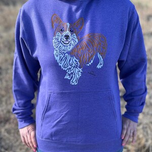 Puede incluir: Una sudadera con capucha morada con un bolsillo delantero y un gráfico de un perro corgi. El diseño del corgi presenta detalles de pelaje marrón y blanco y un patrón decorativo azul claro. La sudadera con capucha está hecha de un material suave.