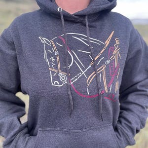 Könnte beinhalten: Dunkelgrauer Kapuzenpullover mit einem weißen, rosa und beigefarbenen Pferdekopf- und Zaumzeug-Design. Die Grafik zeigt eine detaillierte Umrisszeichnung eines Pferdekopfes und -geschirrs mit einer rosa Trense und beigen Akzenten. Der Hoodie hat eine Kapuze mit Kordelzug und eine Vordertasche.