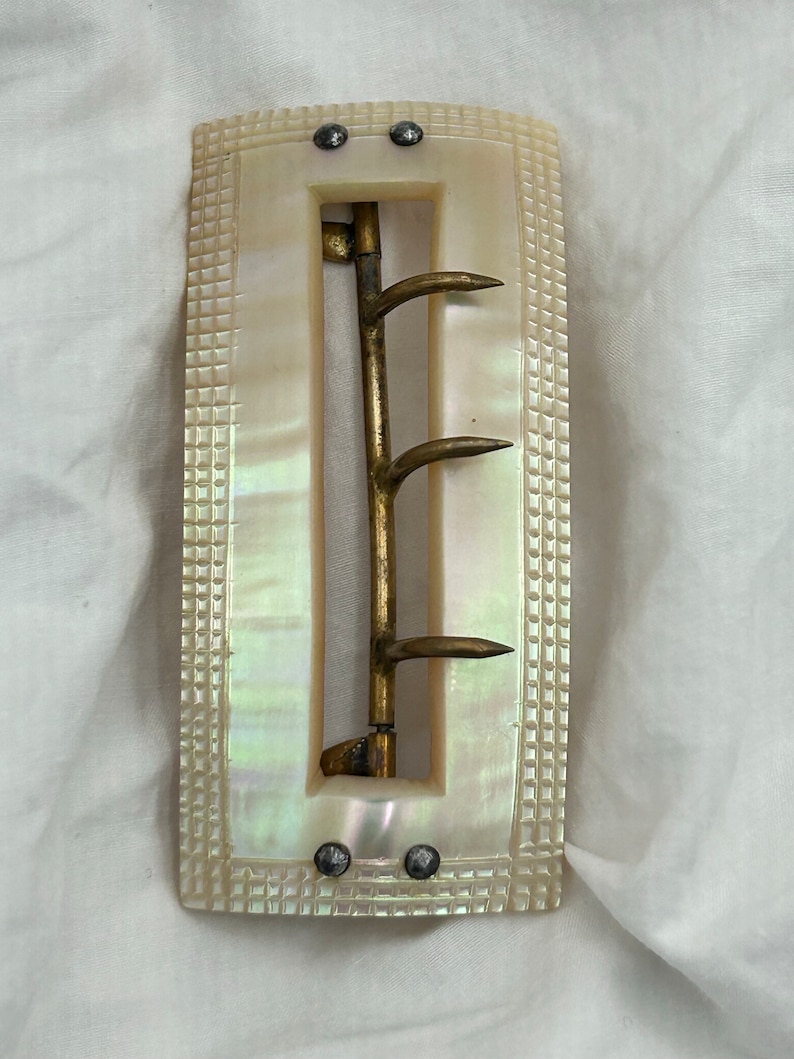 Puede incluir: Una hebilla de cintur&oacute;n vintage de n&aacute;car con un gancho de lat&oacute;n y tres puntas. La hebilla es rectangular y tiene una superficie texturizada.
