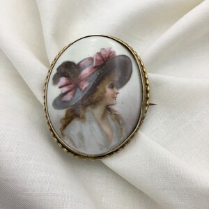 Pu&ograve; includere: Broche ovale in porcellana con cornice dorata. La broche presenta un ritratto dipinto di una donna con un grande cappello a tesa larga con un nastro rosa.