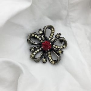 May include: A vintage flower brooch with a silver-tone metal finish. The brooch features a central red gemstone surrounded by clear, small crystals and curved petals. A classic accessory for adding a touch of elegance.