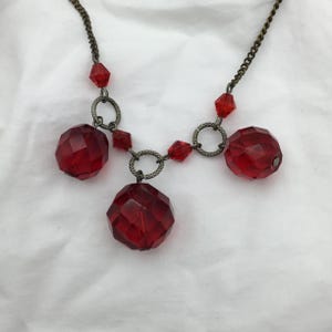 May include: A vintage necklace featuring a dark chain and decorative red faceted beads. The necklace has three large, round beads and smaller diamond-shaped beads. The metal has a dark, antique finish.