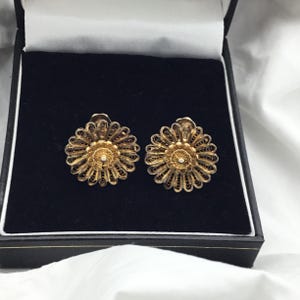 以下が含まれることがあります： 黒い宝石箱に入った、金色のフィリグリーフラワーイヤリングのペア。各イヤリングは、デイジーに似た繊細で渦巻く金属細工に囲まれた、中央の真珠のようなビーズが特徴です。イヤリングは、濃い青色のベルベットの裏地に対して表示されます。